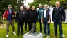A group photo of seven men standing outdoors around a small sculpture of a dragon riding a scooter.
