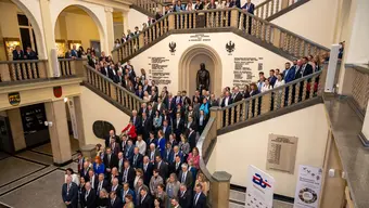 Zdjęcie grupowe dużej liczby osób ustawionych na reprezentacyjnych schodach wewnątrz zabytkowego budynku uczelni; w tle widoczny pomnik, napisy i herby.