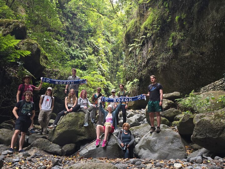 A group of people posing for a photo standing and sitting on rocks in the middle of a forrest. Some are holding scarfs of the Bozon club.