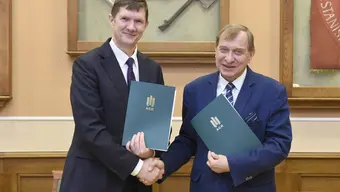 Conference room in the A0 building. From the left, dressed in suits, is the WSB-NLU Rector and Professor Jerzy Lis, AGH UST Rector. They are holding dark green folders with documents in their left hands. They shake hands in a congratulatory gesture. Behind them, there are glass cases with the AGH UST emblem and insignia: a sceptre and ceremonial axe.