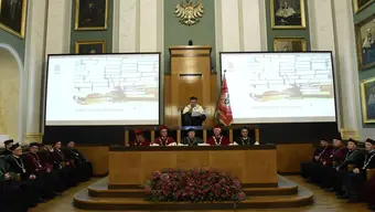 Zdjęcie z uroczystości Dnia Edukacji Narodowej, wykonane w auli. Rektor przemawia, stojąc, przed nim w stallach siedzą Prorektorzy, obok w stallach - Dziekani.