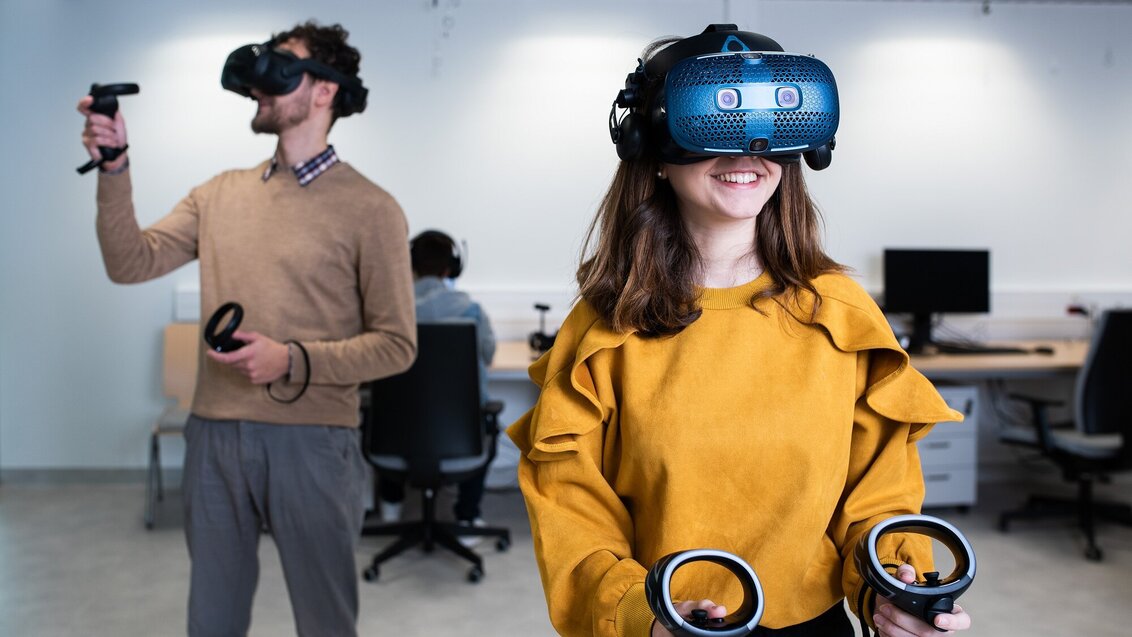 Two students wearing VR headsets and holding controllers in a spacious room. Another student is sitting at a desk in the back of the room.