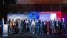 Grupa ok. 30 osób stojących na scenie, uczestników konferencji, za nimi baner z grafiką wydarzenia
