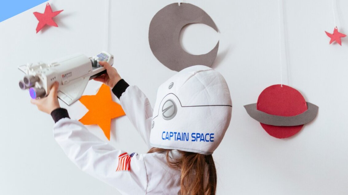 Dziewczynka w białym kombinezonie astronauty, stoi tyłem do obiektywu aparatu. Trzyma uniesioną do góry zabawkę, rakietę. Na ścianie przed nią wiszą wycięte z papieru, kolorowe gwiazdy i planety. 