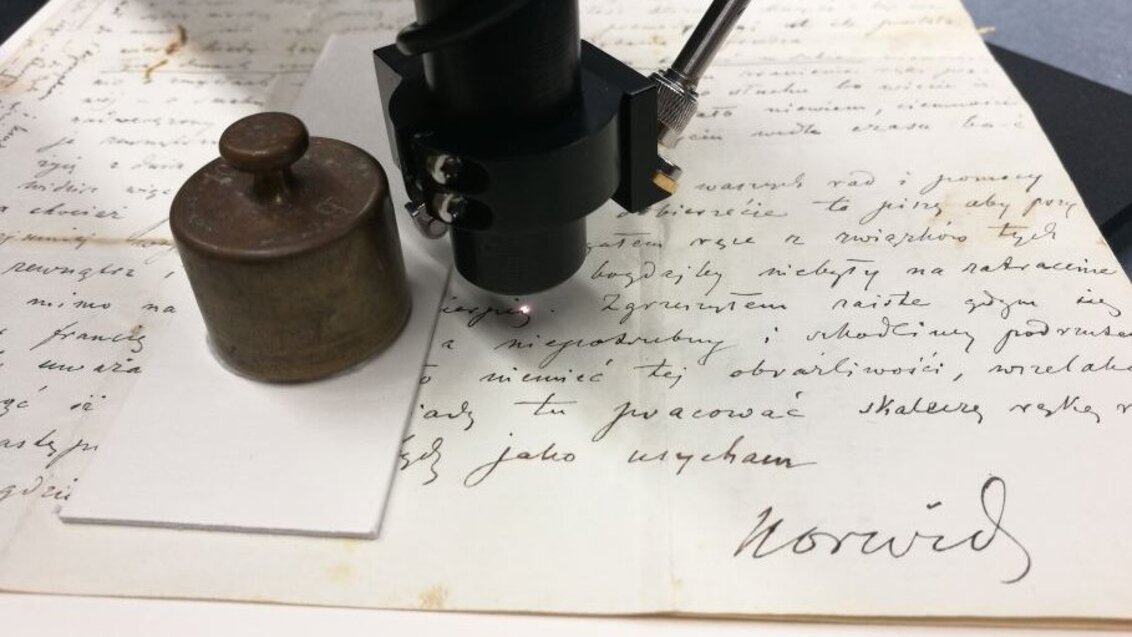 Image of a letter by Norwid being examined with a specialistic device. A weight on the left side.