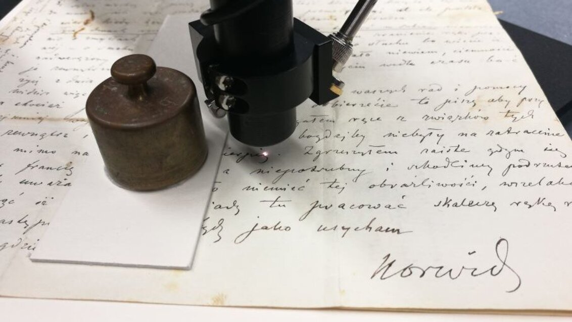 Zdjęcie listu Cypriana Kamila Norwida. Nad nim fragment specjalistycznego sprzętu do badań przypominający soczewkę mikroskopu.