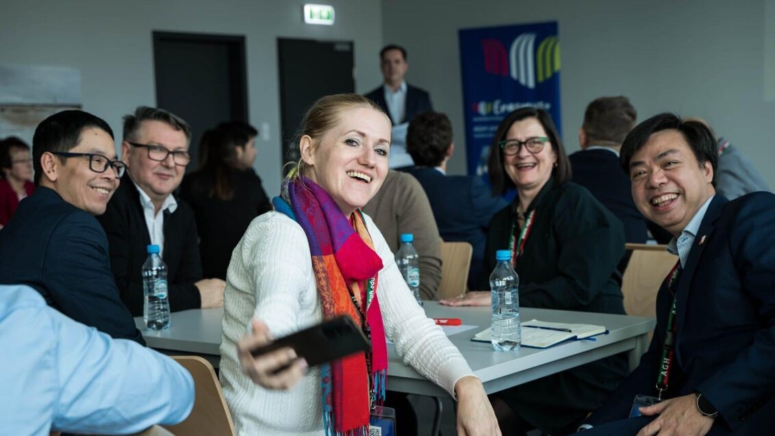 An image of a group of people sitting at a table taking a selfie. In the background, there is a banner of the AGH University Erasmus+ Staff Week