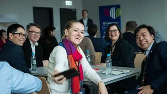 Na zdjęciu grupa uśmiechniętych ludzi. Siedzą przy stole, w pomieszczeniu. Jedna z osób - kobieta - trzyma w dłoni telefon, którym robi zdjęcie. Pozostałe osoby patrzą w kierunku aparatu.