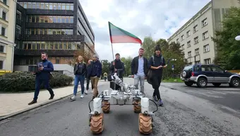 The rover Kalman driving on one of the AGH University campus’ inner roads. Behind it, there are 7 people, students, following it. One of the female students holds an AGH University flag in her hands.
