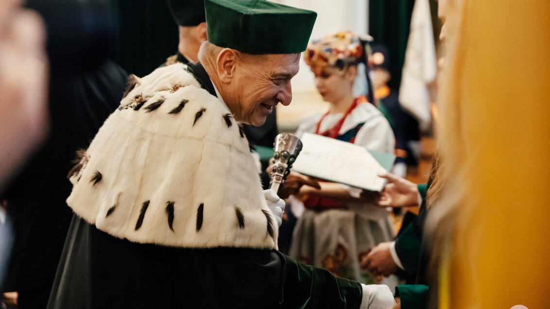 Immatrykulacja. Rektor Jerzy Lis stoi bokiem do obiektywu, ubrany jest w odświętną togę w zielonym kolorze oraz zielony biret, na ramionach ma biały kołnierz. W lewej dłoni trzyma berło zdobione kolorowymi kamieniami, prawą dłonią uścisnął rękę studentki w geście gratulacji.