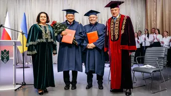 Four university representatives in academic ceremonial attire stand on a stage during an honorary doctorate ceremony. Professors Jerzy Lis and Alojzy Nowak hold diploma folders, one carrying a bouquet of red roses. The Rector and former Rector of the Carpathian National University stand to their sides. Polish and Ukrainian flags are visible beside a podium, while attendees stand in the background.
