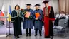 Four university representatives in academic ceremonial attire stand on a stage during an honorary doctorate ceremony. Professors Jerzy Lis and Alojzy Nowak hold diploma folders, one carrying a bouquet of red roses. The Rector and former Rector of the Carpathian National University stand to their sides. Polish and Ukrainian flags are visible beside a podium, while attendees stand in the background.