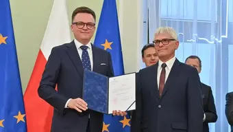 Na zdjęciu Marszałek Sejmu Szymon Hołownia (po lewej) trzyma w dłoniach akt powołania. Obok niego stoi dr. Leszek Rymarowicz. W tle widać kilka innych osób oraz flagi Polski i UE.