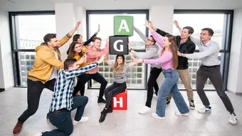 Grupa młodych osób stoi w pomieszczeniu z dużymi oknami, pozując wokół trzech dużych, kolorowych puf ustawionych jeden na drugim. Na pufach widnieją litery „A” (zielony puf), „G” (czarny puf) i „H” (czerwony puf).