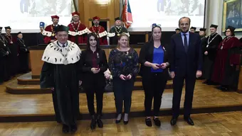 Na zdjęciu trzy odznaczone pracownice, które stoją obok siebie. Po lewej stronie Rektor, po prawej wicewojewoda. Za stojącymi osobami stalle, w których siedzą prorektorzy i dziekani. Zdjęcie wykonane w auli AGH.