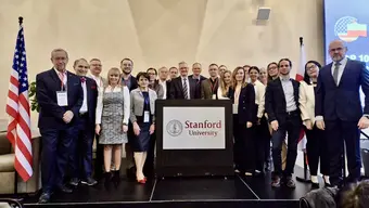 A large group of adults standing on a stage behind a podium labeled “Stanford University,” with U.S. and Polish flags.