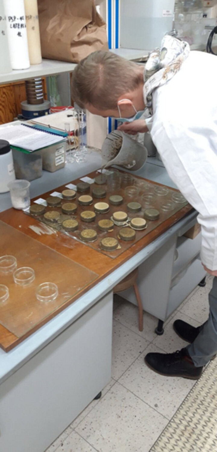 Na zdjęciu widać naukowca w laboratorium, który pochyla się nad stołem z okrągłymi próbkami. Na stole widać kilkanaście próbek. Naukowiec trzyma w ręku pojemnik wypełniony przygotowanym wcześniej zaczynem uszczelniającym i wlewa do próbek