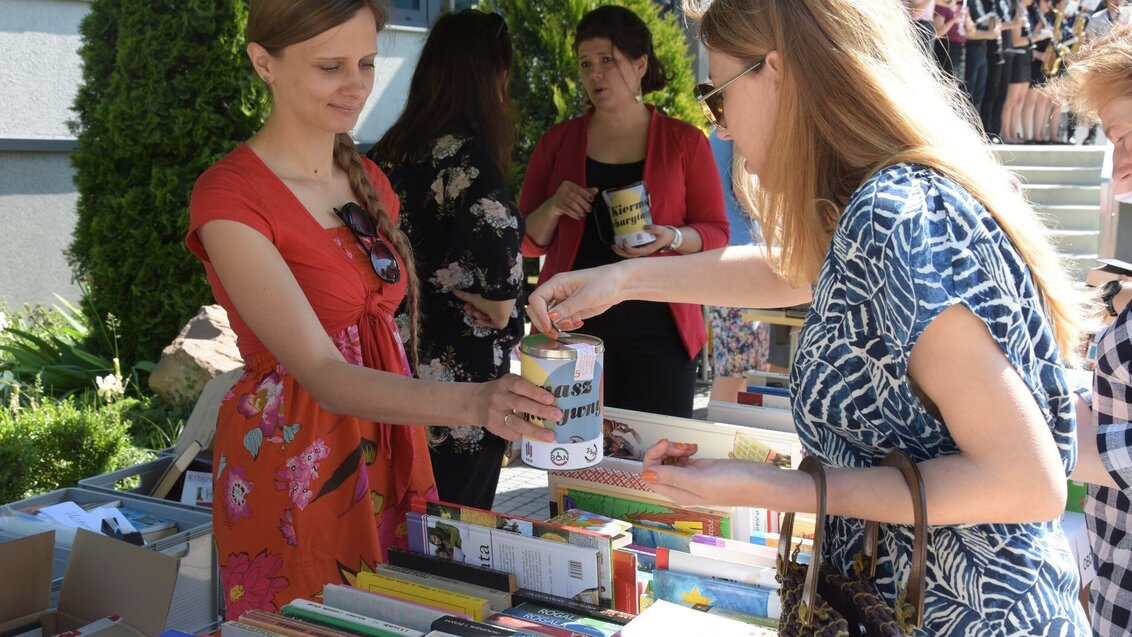 Na zdjęciu dwie kobiety stoją po przeciwnych stronach stolika z książkami. Jedna z nich płaci, druga trzyma w dłoni puszkę na datki. Zdjęcie wykonane przed budynkiem Biblioteki Głównej AGH.