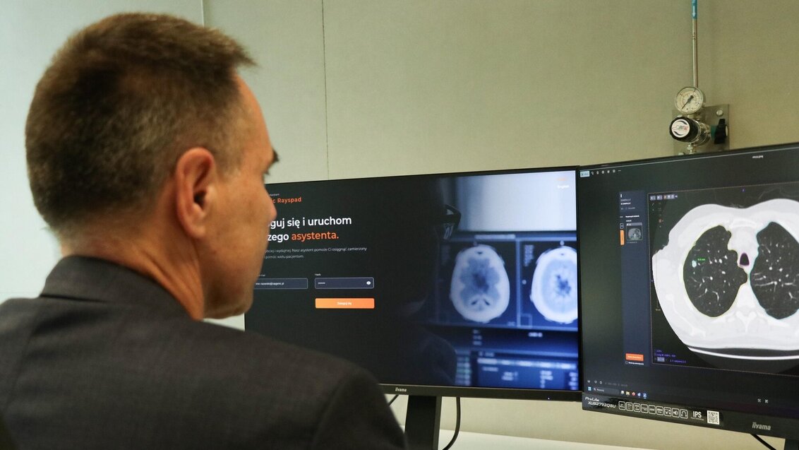 A suited professional reviews brain scans and a chest CT on dual monitors, using medical software in Polish—suggesting diagnostic analysis.