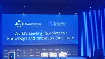 Zdjęcie sceny konferencyjnej z wyświetlonym slajdem z nazwą wydarzenia. Na scenie trzy białe fotele.