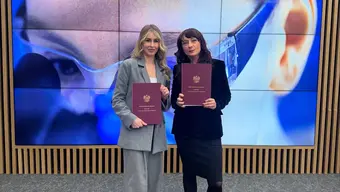 An image of the AGH University scientists posing with burgundy folders with the Polish eagle, stading in front of a screen displaying a hoto of a female scientist in a laboratory