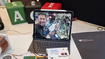 An image of a tablet at an exhibition stand displaying a photo of Sławosz Uznański-Wiśniewski on the ISS wearing the AGH University-designed MXene wristband. An AstroPi device is laid on the table. In the background, there are three green, black, and red poufs.