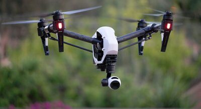 Dron z kamerą zawieszony w powietrzu na tle rozmytej zieleni.