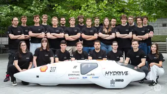 The image features a team in black shirts around a futuristic vehicle labeled "HYDRIVE," with sponsor logos and the number "217". The setting is outdoors.