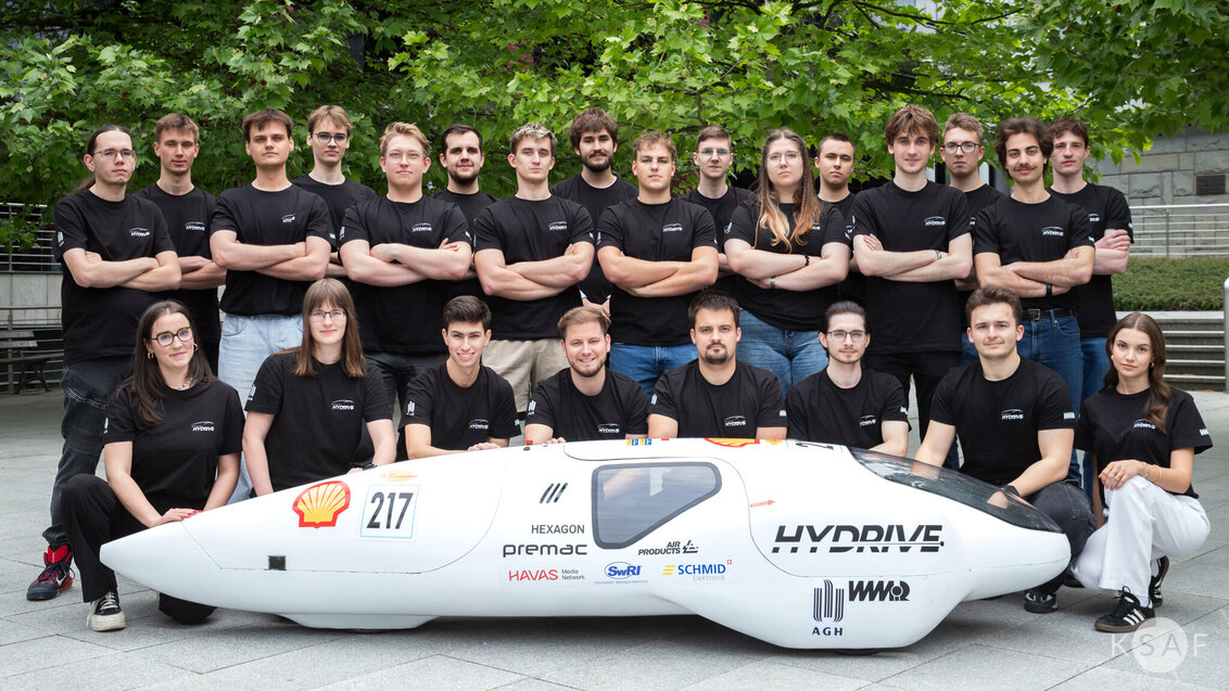 The image features a team in black shirts around a futuristic vehicle labeled "HYDRIVE," with sponsor logos and the number "217". The setting is outdoors.