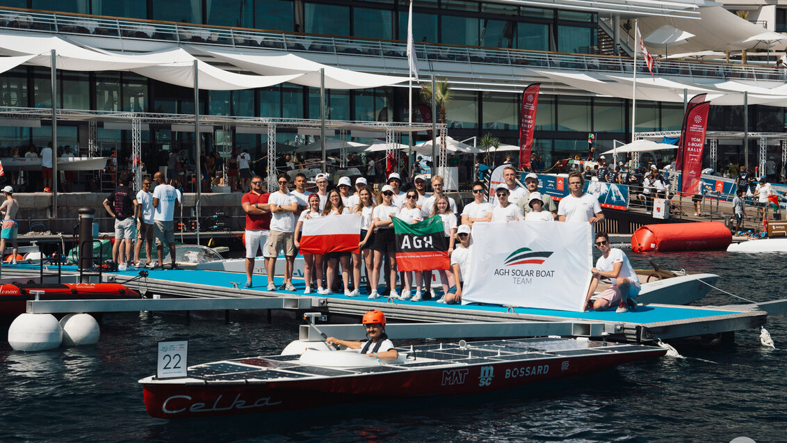 Grupowe zdjęcie członków AGH Solar Boat wykonane w basenie jachtowym portu. Studenci stoją na pomoście, przed nimi, na wodzie Celka. W łodzi siedzi sternik. W tle widoczne budynki portu wzdłuż linii brzegowej