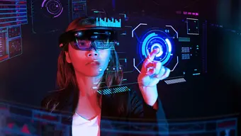 Colourful image with graphic elements. A woman in VR goggles in a virtual room points her finger to a holographic screen hovering in front of her.
