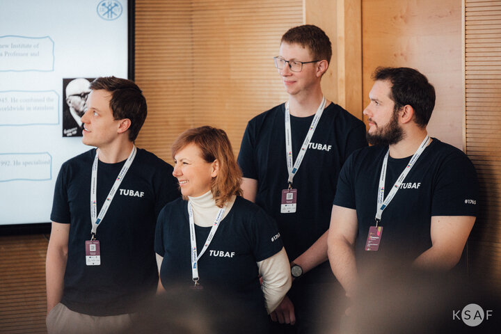 Zdjęcie grupy osób stojących obok ekranu z wyświetlaną prezentacją. Ubrani w takie same koszulki z napisem "TU Bergakademie Freiberg". Każda osoba ma zawieszony na szyi identyfikator z nazwą wydarzenia.