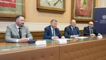 Czterech mężczyzn w garniturach siedzi przy długim stole konferencyjnym w eleganckiej sali z historycznymi sztandarami w gablotach na ścianach. Na stole znajdują się tabliczki z nazwiskami, dokumenty oraz długopisy.