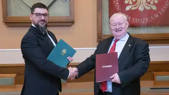An image of two men in business attire, Rector Wiśniowski and Jakub Progorowicz, shaking hands after having exchanged folders of their respective entities.