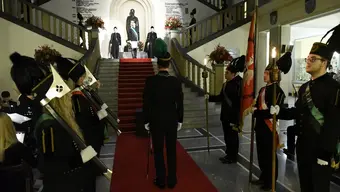 Zdjęcie inscenizacyjne tradycyjnego "skoku przez skórę" wykonane w holu budynku głównego AGH. Na zdjęciu studenci i studentki ubrani w odświętne górnicze mundury, trzymają w dłoniach sztandary i paradne topory górnicze. Światło jest przytłumione, a reflektor skierowany na schody, po których schodzą studenci i studentki, którzy będą wykonywać skok i składać przysięgę górniczą.