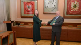 Image of the parties to the agreement shaking hands, holding the documents in a room with decorative banners hanging on a wall