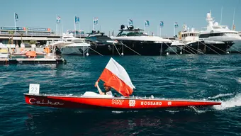 Zdjęcie Celki na morzu. Sternik trzyma w ręce flagę Polski. W tle widoczne cumujące w porcie statki.