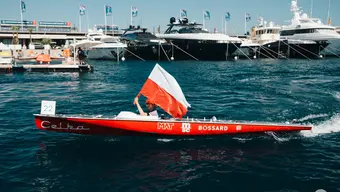 Zdjęcie Celki na morzu. Sternik trzyma w ręce flagę Polski. W tle widoczne cumujące w porcie statki.