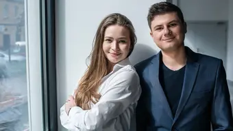 Dwie osoby pozują do zdjęcia, stojąc obok siebie przy dużym oknie w korytarzu budynku. Osoba po lewej (kobieta) ubrana jest w białą koszule, po prawej (mężczyzna) - w granatową marynarkę.