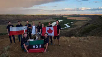 Grupowe zdjęcie zespołu AGH Space Systems na tle kanadyjskiego krajobrazu z rozległą, otwartą przestrzenią i widoczną doliną rzeki. Studenci trzymają w dłoniach rozpostarte flagi Polski, Kanady oraz AGH. Pomiędzy studentami widoczny łazik Kalman.