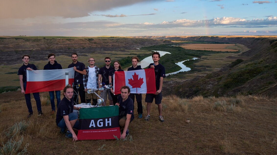 Grupowe zdjęcie zespołu AGH Space Systems na tle kanadyjskiego krajobrazu z rozległą, otwartą przestrzenią i widoczną doliną rzeki. Studenci trzymają w dłoniach rozpostarte flagi Polski, Kanady oraz AGH. Pomiędzy studentami widoczny łazik Kalman.