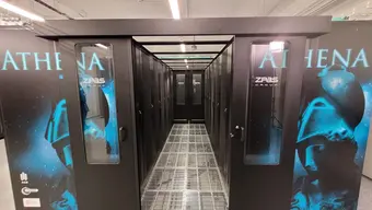 A corridor between two rows of supercomputer modules. It is confined on both sides by black glass doors. The floor is made of a metal grate. The external sides of the modules show a light blue image of a woman with an ancient helmet on. Above the figure, there is an inscription that reads ‘ATHENA’.
