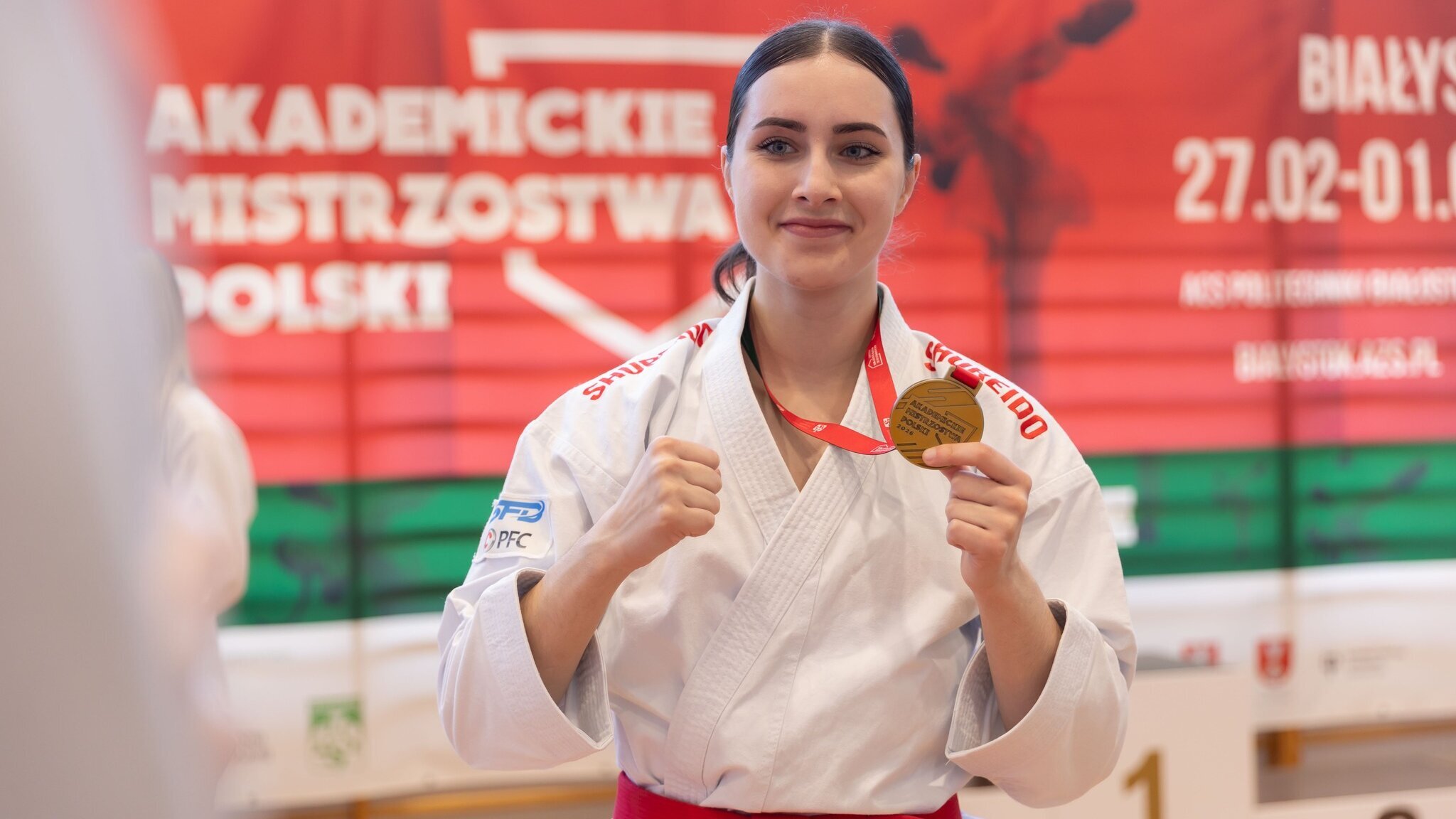 A young woman in a karate uniform with a gold medal on her neck. She is proudly holding it in her hand.