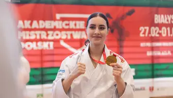 Na zdjęciu zawodniczka karate w kimonie. Na jej szyi wisi złoty medal, który z dumą prezentuje, trzymając krążek w dłoni.