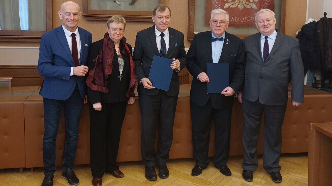 Five people standing in a row (four men and a woman). All of them dressed smartly, two men in the middle hold dark blue document folders.