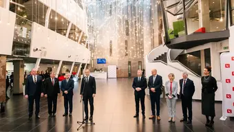 Grupowe zdjęcie osób uczestniczących w spotkaniu wykonane w holu siedziby Krakowskiego Parku Technologicznego. Osoby stoją obok siebie, wśród nich prezydent, który mówi coś do stojącego przed nim mikrofonu.