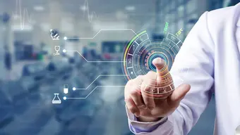 Na zdjęciu ramię w lekarskim kitlu - dłoń z palcem wskazującym na graficzny element w postaci tarczy. W rozmazanym tle pomieszczeni, zapewne laboratoryjne.