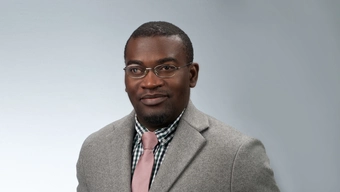 A portrait photo of a young black man wearing glasses, a light suit, a checkered shirt and a pink tie.