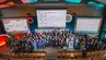 A group photograph of conference participants on stage in an auditorium taken from above.