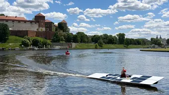 Solarna Delta płynie po Wiśle w pobliżu Wawelu. Na pokładzie jedna osoba w kasku unosi w górę rękę.